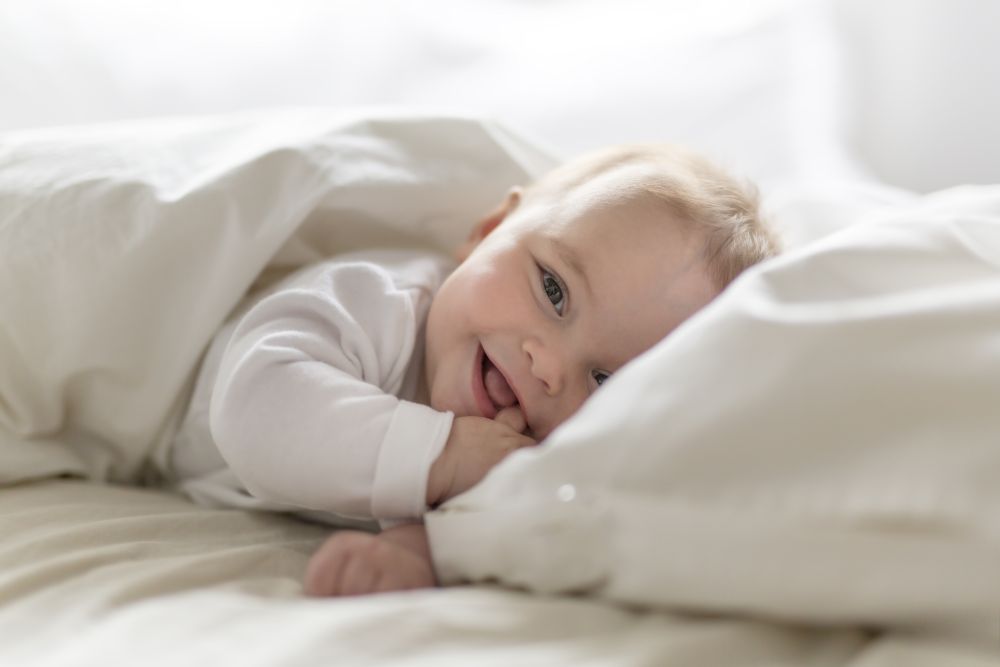 En baby ligger under en dyne og smiler med hånden i munnen.