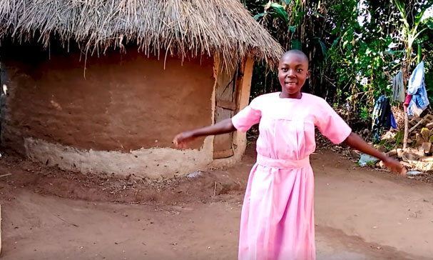 Jente i rosa kjole står foran sin landsby å smiler til kamera