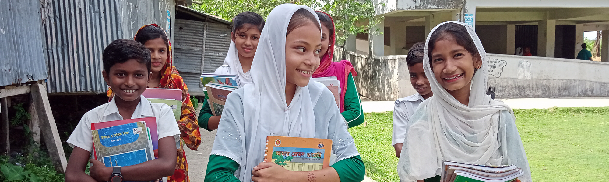 to jenter fra Bangladesh går smilede foran i en gruppe barn. Alle bærer skolebøker med seg.