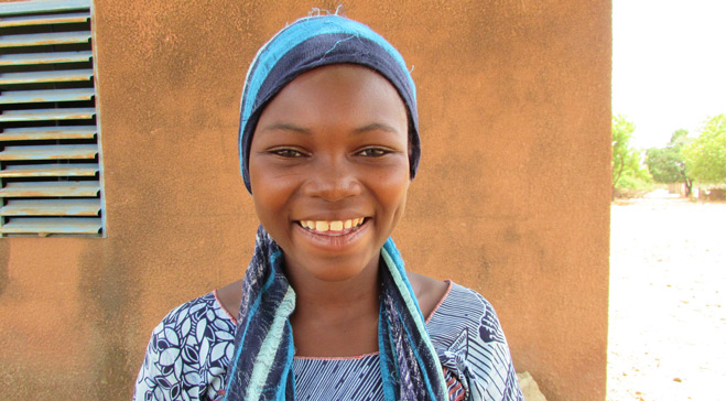 Jente smiler mot kamera utenfor hus i Burkina Faso