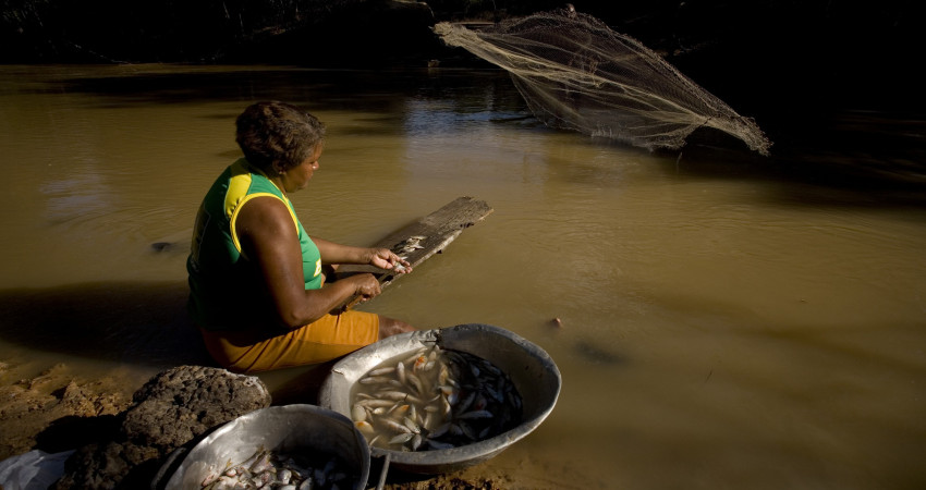 En kvinne sitter på en sten ved elven og renser fisk hun har samlet i to fat