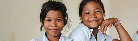 To jenter sitter ved siden av hverandre i et klasserom, begge smilende og glade. De er kledd i hvite skjorter og ser rett mot kameraet.