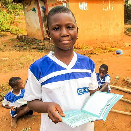 Gutt i Benin viser frem bok utendørs med to klassekamret i bakgrunnen.