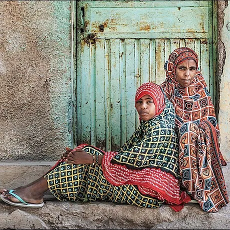 Mor og datter i Etiopia sitter forran grønn-turkis dørr i fargeglade og mønstrete klær.