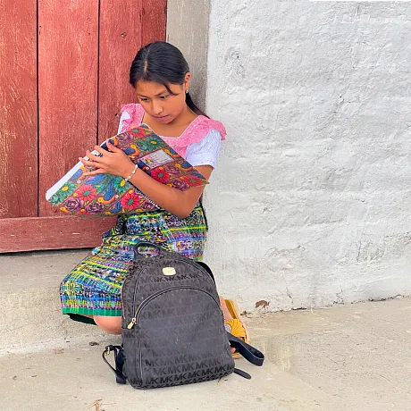 Jente i Guatemala sitter utenfor en dør med en skolesekk og skriver i en notatbok.