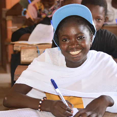 Jente smiler i klasserom og skriver i notatbok.