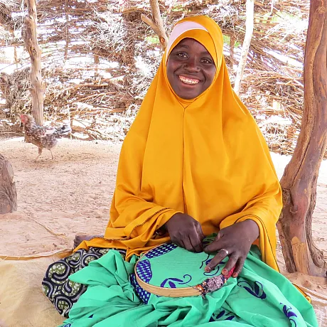 Jente i Niger sitter på bakken og smiler og syr.