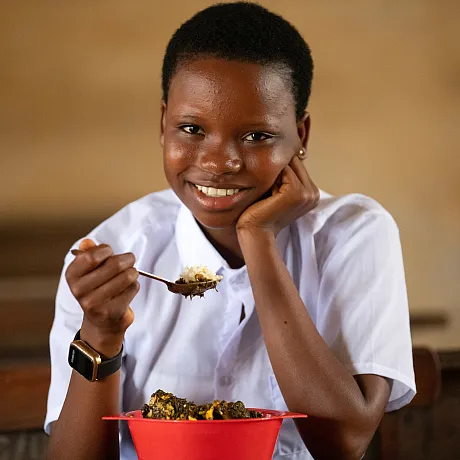 Jente smiler mot kamera når hun løfter opp en skje med mat på skolen.