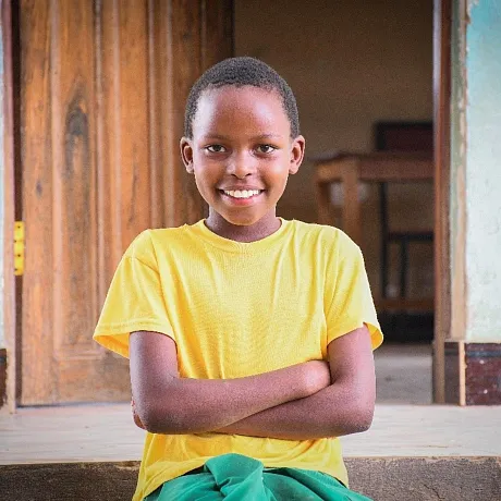 Jente i gul t-skjorte krysser armene forran klasserommet i Tanzania.