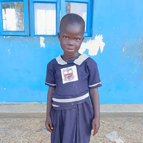 Ung jente i blå skoleuniform står foran skolen i Uganda.